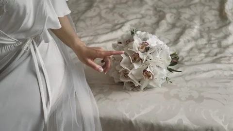 A bride in a white dress sits on the bed and touches her bouquet in close-up Stock Footage 213633488