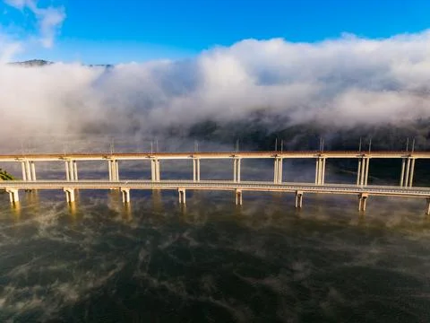 Bridge spans water under cloudy sky in morning light with mist hovering ove.. Stock Photos