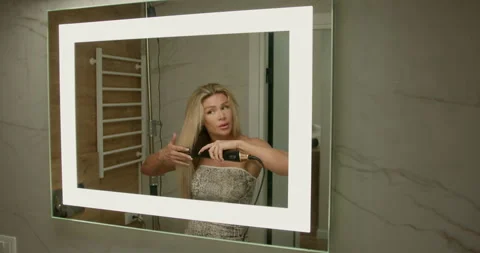 In bright bathroom blonde girl is using hand dryer dry her hair while looking Stock Footage 306123363