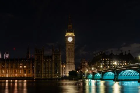 Bright clock face rises above the river and bridge at night, combining urba.. Stock Photos