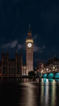 Bright clock face rises above the river and bridge at night, combining urba.. Fotos de archivo