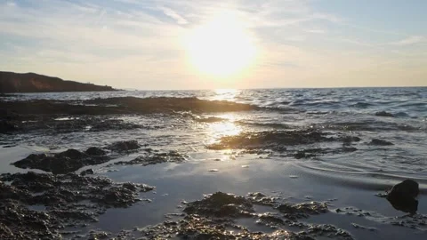 Bright golden sun sets low over calm rocky ocean shore Stock Footage 337410897