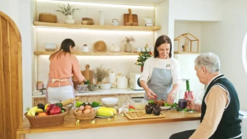 Bright home kitchen where two women prepare a fresh meal while an older man.. Video stock 347061060
