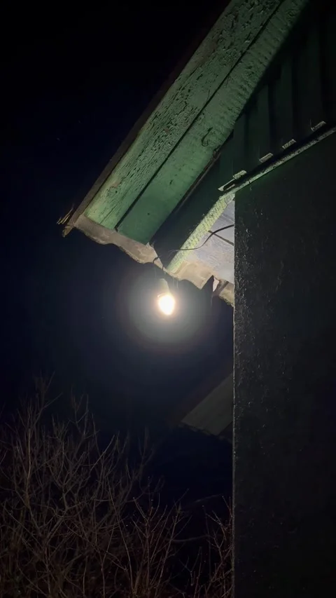 Bright light illuminates the corner of a rustic building's roof at night Stock Footage 319299495