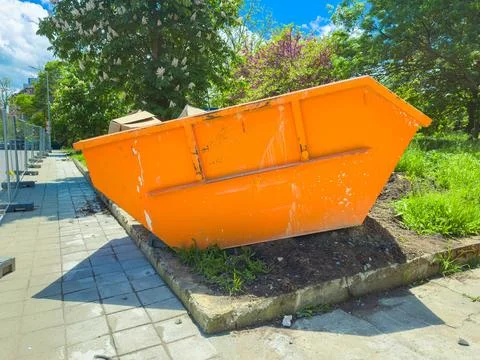Bright orange dumpster stands on the side of a sidewalk with greenery and t.. Stock Photos
