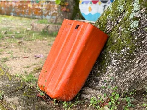 Bright Orange Plastic Container Leaning Against a Mossy Tree Trunk in a Park Stock Photos