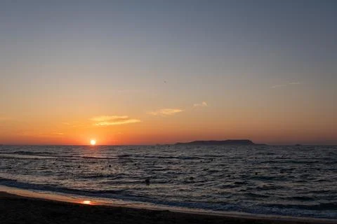 Bright orange sun sets over rippling sea near distant island silhouette Stock Photos