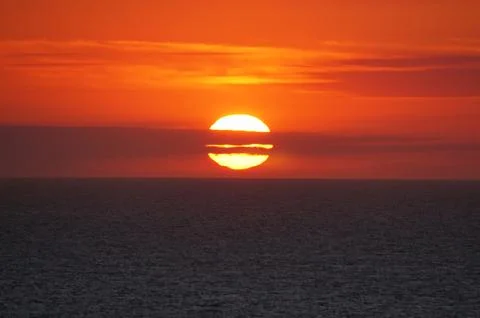 Bright orange sun setting over ocean with dramatic clouds and warm sky grad.. 写真素材
