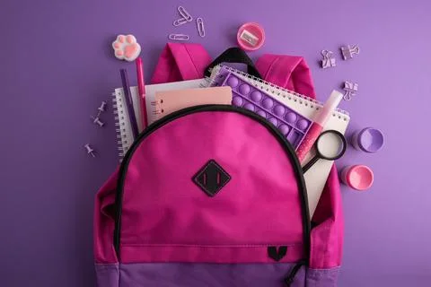 Bright Pink Backpack Filled With School Supplies And Stationery On A Purple.. Stock Photos