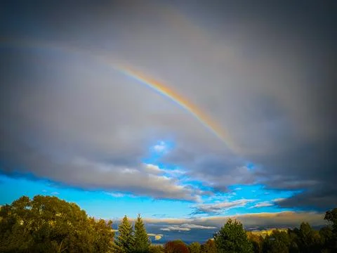Bright rainbow arching through dramatic storm clouds above a colorful tree-.. Фото