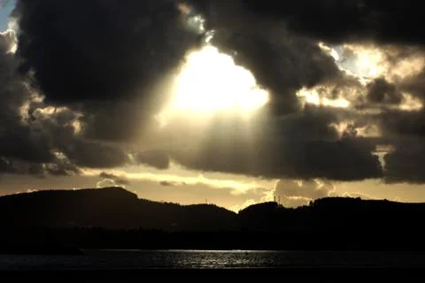 Bright rays on the sun shine through dark clouds over lake Stock Photos