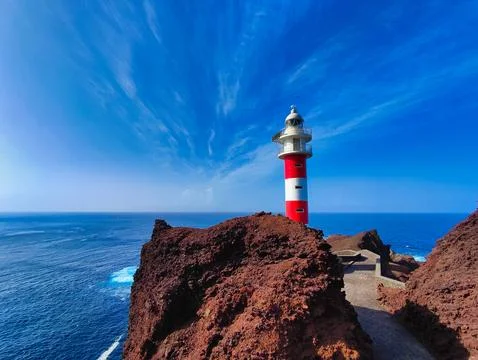 Bright red and white lighthouse stands on rocky cliffs by the ocean under b.. Stock Photos