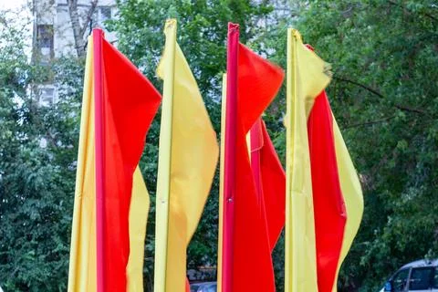 Bright red and yellow flags waving on poles in front of green trees Stock Photos