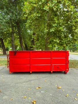A bright red dumpster is parked on a gray pavement in a park. Tall green tree Stock Photos