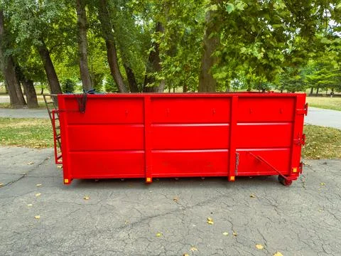 A bright red dumpster is placed on a paved surface in a park. Green trees pro Stock Photos