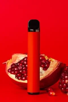 Bright red vape device stands beside cut pomegranate on red background Stock Photos