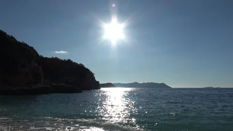 Bright sun with rays reflecting on the sea and dark rocky cliffs under blue  Vidéo 332860927