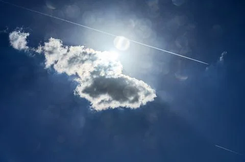 Bright sun shines through a single cloud with contrails in a blue sky Stock Photos