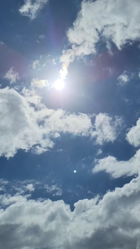 Bright sun shining through white clouds in blue sky, vertical POV 스톡 동영상 330082377