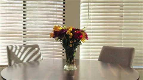 Bright sunlight shines through blinds, casting soft patterns on a vase of fresh Stock Footage 304857967