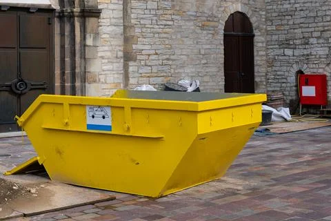 Bright yellow construction dumpster positioned adjacent to stone building Stock Photos