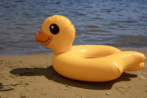Bright yellow inflatable duck float resting on sandy beach by water Stock Photos