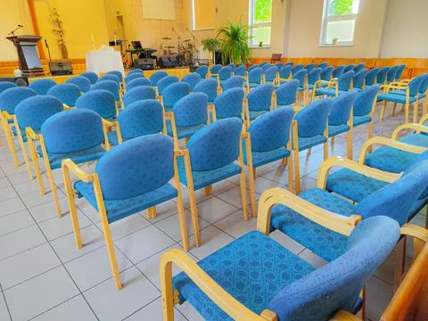 Brightly lit hall with rows of empty blue chairs facing a stage. Stock Photos