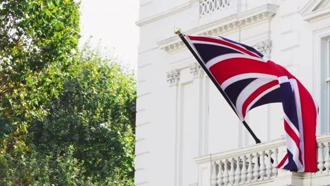 British flag waving in the breeze outside a white building with greenery nearby Stock Footage 331801812