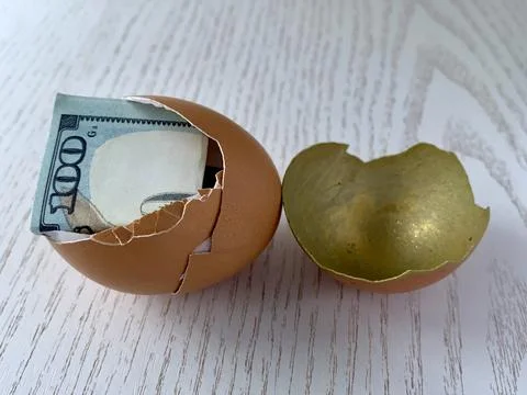 A broken egg shell shows a rolled-up banknote nestled inside. The golden Foto stock