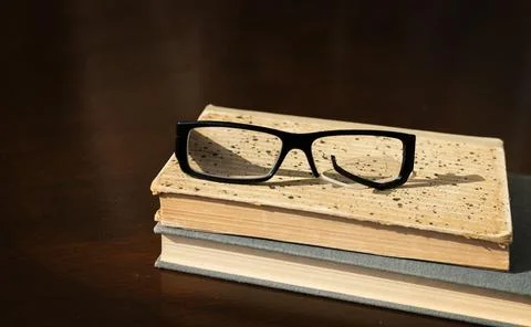 A broken eyeglass frame lies on a stack of old books on a brown wooden table Stock Photos