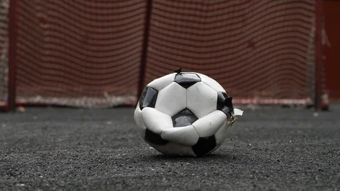 Broken half deflated soccer ball lie on black rubber field against blurred gate