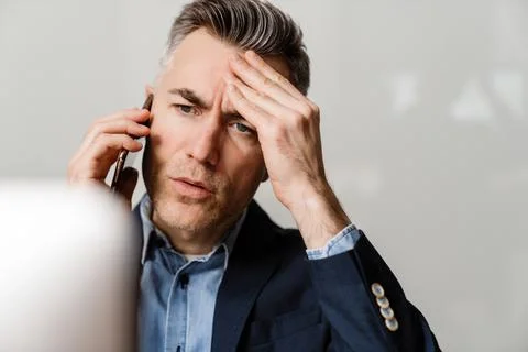 Brooding grey man talking on mobile phone while working with computer Stock Photos