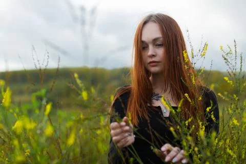 A brooding red-haired girl with a sad face stands among the wildflowers Stock Photos