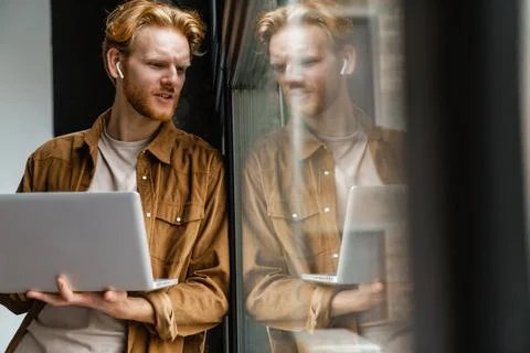 Brooding redhead guy in wireless earphones working with laptop Stock Photos