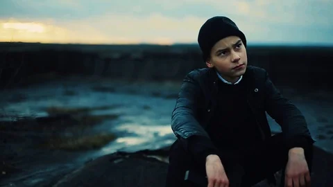 Brooding sad boy with gun in hand sitting on stone. Portrait of depressed guy Stock Footage 98603495