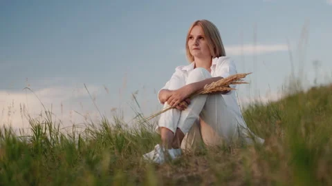 A brooding woman in a white suit sits on a hill among the grass in a picturesque Stock Footage 136219707