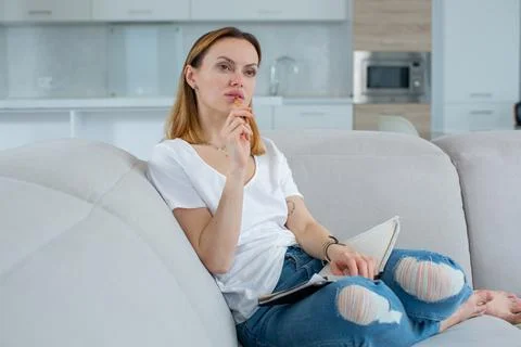 A brooding young girl writes notes in a notepad, sitting at home on the couch Stock Photos