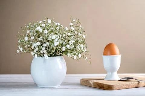 Brown egg in egg cup and gypsophila in white vase on light background. Easter Stock Photos