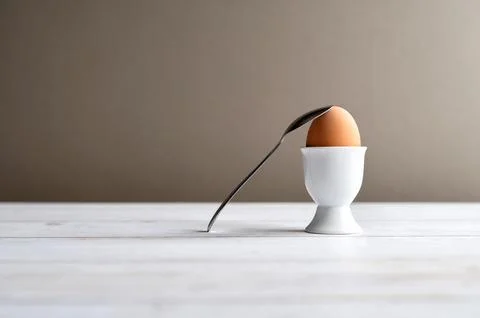 Brown egg in a white ceramic egg cup and spoon on a light wooden surface with Stock Photos