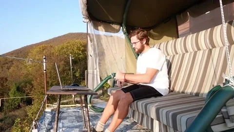 A brown-haired bearded man is sitting on the balcony while working using his lap Stock Footage 235886438