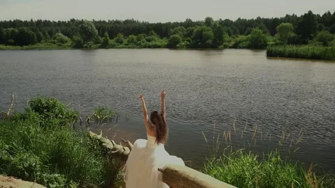 A brown-haired bride in white wedding dress sits on trunk of tree above water Stock Footage 151109090