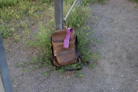 Brown leather backpack with purple strap rests on the ground near a metal pol Stock Photos