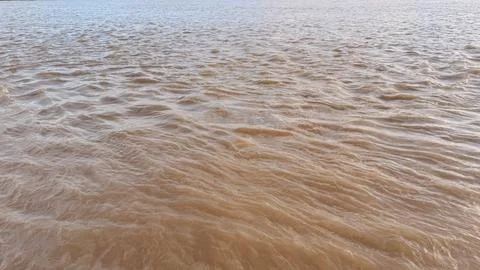 Brown murky river water flows with small choppy waves across the wide expanse Stock Photos
