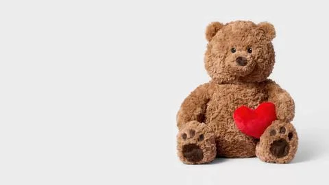A brown stuffed bear sits on a copy space background while holding a red heart Fotos de archivo