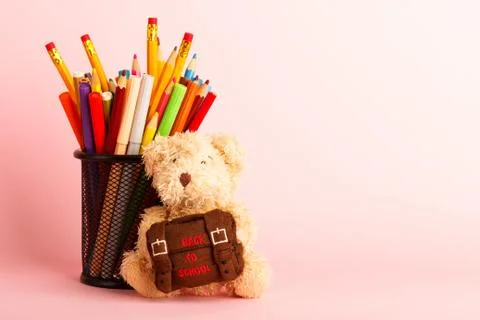 Brown teddy bear with school backpack and glass with felt-tip-pens, pink back 库存照片
