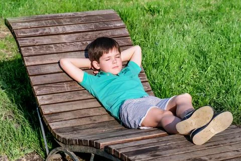 Brunet boy of 8 years in T-shirt and shorts lies on wooden deck chair in the  Stock Photos