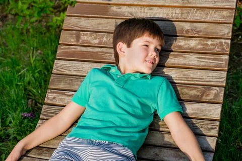 Brunet boy of 8 years in T-shirt lies on wooden deck chair in the sun against Stock Photos