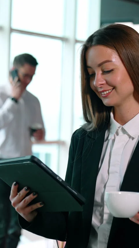 Brunette in black jacket stands looking at I-pad and holding a cup in hand. Man Stock Footage 325040440