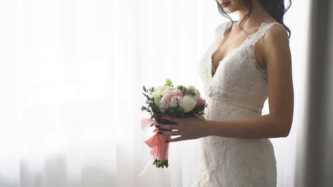 A brunette bride stands at the window and sniffing the Bridal bouquet Stock Footage 76941710