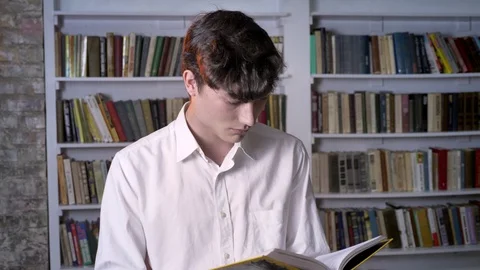 Brunette man is reading book, watching at camera, smiling, library on background Video stock 92467256